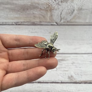 May include: A detailed bronze-colored bee figurine with intricate wing and body details. The bee is posed with its wings slightly raised, showcasing the delicate veining. The figurine is a decorative item, possibly a brooch or charm.