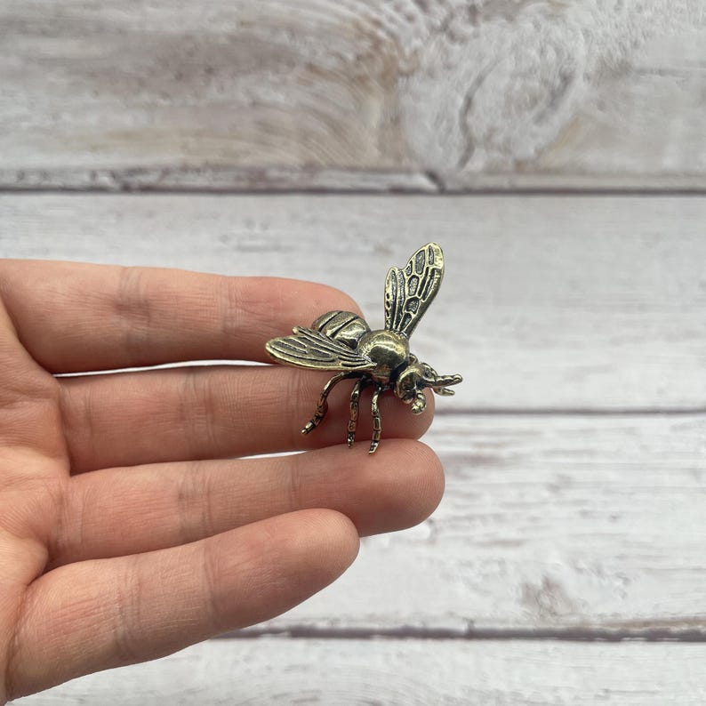 May include: A detailed bronze-colored bee figurine with intricate wing and body details. The bee is posed with its wings slightly raised, showcasing the delicate veining. The figurine is a decorative item, possibly a brooch or charm.