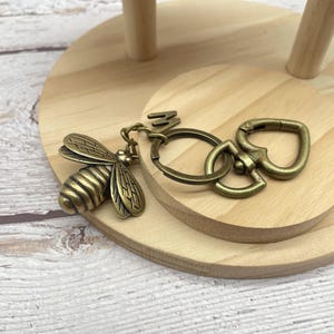 Pu&ograve; includere: Un portachiavi a forma di ape color bronzo con un ciondolo a forma di ape dettagliato, un portachiavi e una chiusura. L'ape ha ali e corpo strutturati. Il portachiavi &egrave; su una superficie di legno chiaro.