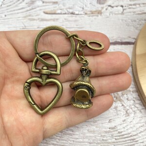 Op de afbeelding: Antieke messingkleurige sleutelhanger met een hartvormige karabijnhaak, een ronde sleutelring en een kleine lantaarnbedel met een slang. De sleutelhanger is van metaal en heeft een vintage uitstraling.