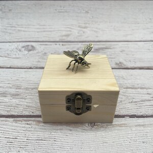 May include: A small, light-colored wooden box with a metal clasp. A detailed, bronze-colored bee figurine rests on top of the box. The box has a simple, rustic design, and the bee adds a decorative touch.