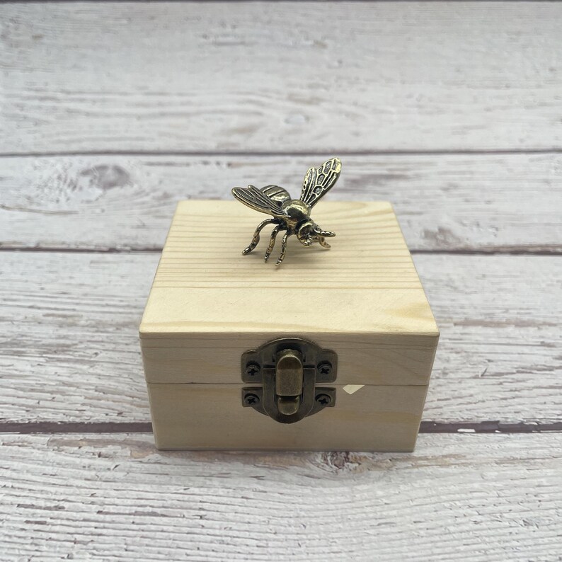 May include: A small, light-colored wooden box with a metal clasp. A detailed, bronze-colored bee figurine rests on top of the box. The box has a simple, rustic design, and the bee adds a decorative touch.