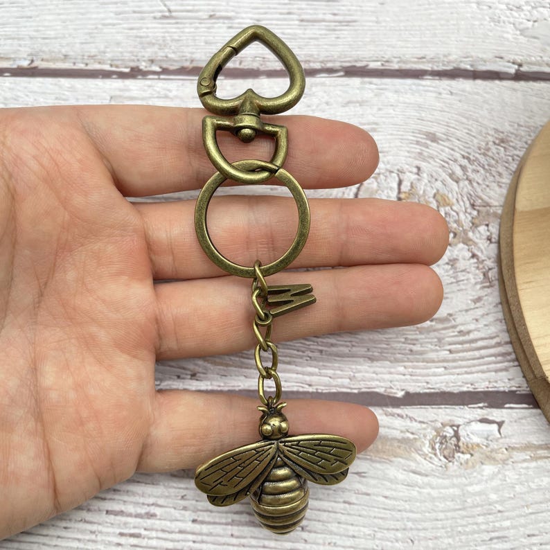 Pu&ograve; includere: Portachiavi color ottone antico con un ciondolo a forma di ape. Il portachiavi ha una chiusura a forma di cuore, un ciondolo a forma di chiave e un portachiavi rotondo. Il ciondolo a forma di ape presenta ali e segmenti del corpo dettagliati.