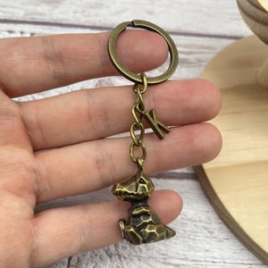 May include: Antique brass-colored keychain featuring a small, textured dinosaur figurine. The keychain includes a chain and a letter "K" charm. The dinosaur is in a sitting position, with detailed scales.