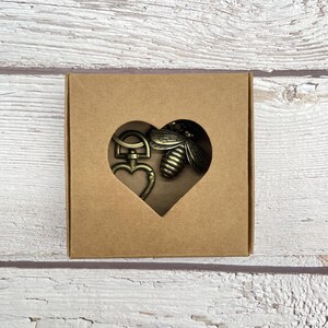 Pu&ograve; includere: Un set di portachiavi color bronzo in un ritaglio a forma di cuore in una scatola di cartone marrone. Il set include una chiusura a forma di cuore e un ciondolo a forma di ape. La scatola &egrave; su una superficie di legno bianco e marrone chiaro.