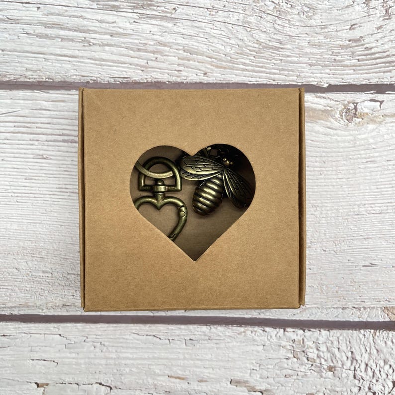 Pu&ograve; includere: Un set di portachiavi color bronzo in un ritaglio a forma di cuore in una scatola di cartone marrone. Il set include una chiusura a forma di cuore e un ciondolo a forma di ape. La scatola &egrave; su una superficie di legno bianco e marrone chiaro.