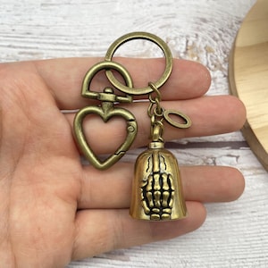 May include: Antique brass-colored keychain featuring a heart-shaped clasp and a bell charm. The bell charm has a skeleton hand design. The keychain includes a key ring and a small, oval-shaped charm.