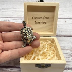 Op de afbeelding: Een kleine, bronskleurige schildpadfiguur wordt voor een open houten kist gehouden. De kist is gevuld met versnipperd papier en heeft de tekst "Custom Your Text Here" gegraveerd. De schildpad heeft gedetailleerde schaal- en ledemaatkenmerken.
