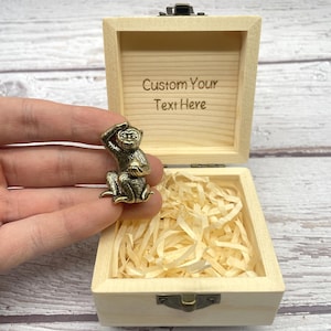 May include: A small, detailed brass monkey figurine held near an open wooden box. The box is filled with shredded paper and has the text "Custom Your Text Here" engraved on the inside of the lid. The monkey is in a seated position, with one hand raised to its head.