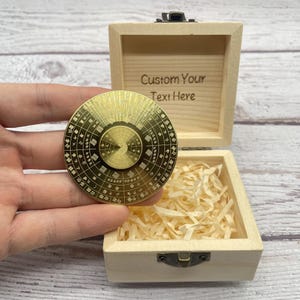 Puede incluir: Un spinner circular dorado con intrincados diseños grabados y texto se sostiene frente a una caja de madera abierta. La caja está llena de papel triturado y tiene el texto "Custom Your Text Here" grabado en el interior de la tapa.