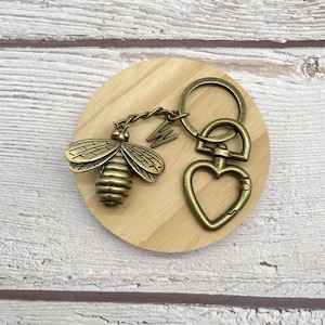 Pu&ograve; includere: Un portachiavi a forma di ape color bronzo con chiusura a forma di cuore e un ciondolo con la lettera "W". L'ape ha ali e segmenti del corpo dettagliati. Il portachiavi &egrave; su un sottobicchiere rotondo di legno.