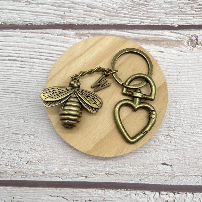 Pu&ograve; includere: Un portachiavi a forma di ape color bronzo con chiusura a forma di cuore e un ciondolo con la lettera "W". L'ape ha ali e segmenti del corpo dettagliati. Il portachiavi &egrave; su un sottobicchiere rotondo di legno.