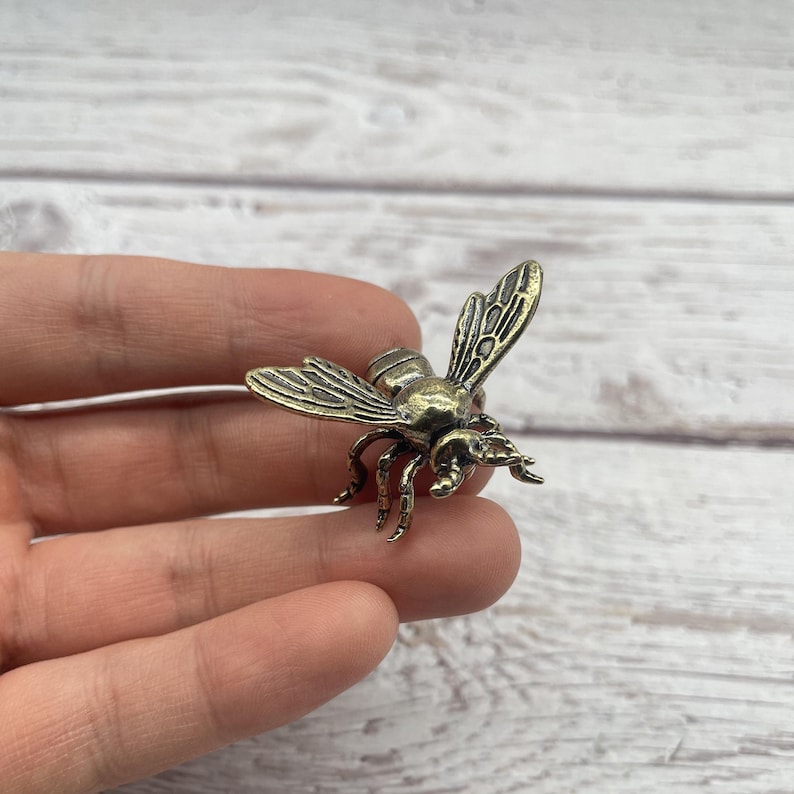 May include: A detailed bronze-colored bee figurine with intricate wing and body details. The bee has six legs and antennae. The figurine is a decorative item, possibly a brooch or charm.