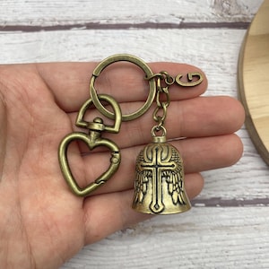 以下が含まれることがあります： アンティーク真鍮色のキーホルダー。ハート型の留め金と、十字架と翼のデザインのベルチャームが特徴です。キーホルダーにはリングと小さなチャームが含まれています。金属はヴィンテージで使い古された外観をしています。