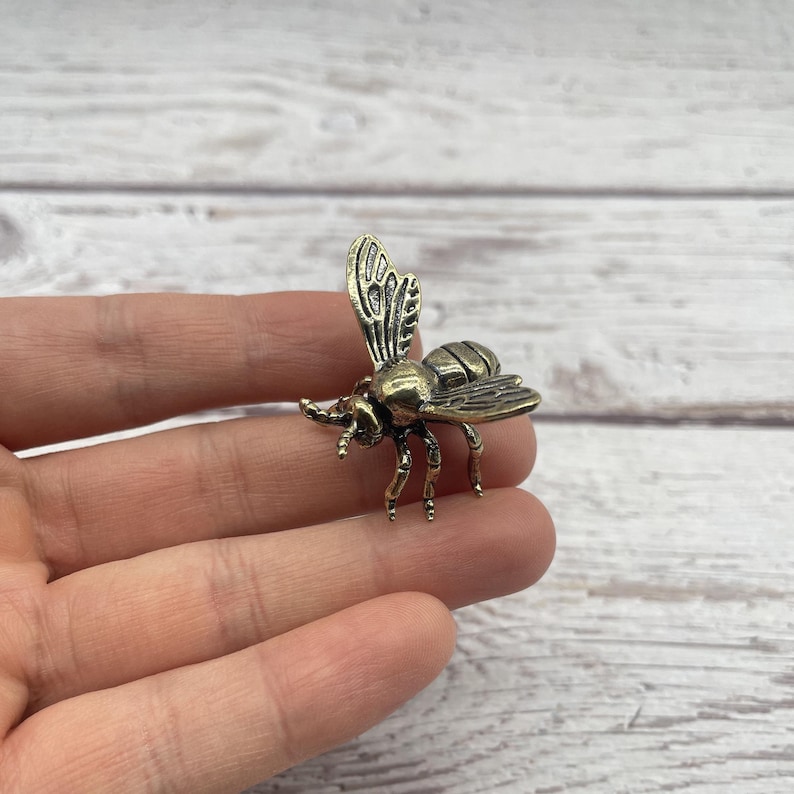 May include: A small, detailed brass bee figurine with outstretched wings and six legs. The bee is a golden-brown color and appears to be made of metal. The wings have intricate vein detailing. The bee is a decorative item.
