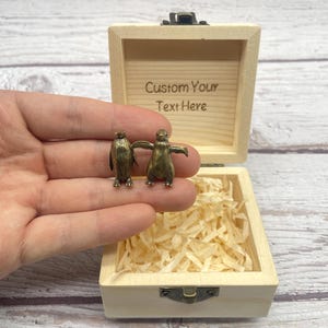 Puede incluir: Dos pequeñas figuras de pingüinos de color bronce se sostienen frente a una caja de madera abierta. La caja está llena de papel triturado y tiene el texto "Custom Your Text Here" grabado en el interior de la tapa.