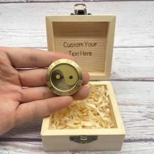 Puede incluir: Un símbolo redondo y dorado de Yin y Yang se sostiene frente a una caja de madera abierta. La caja contiene papel triturado y tiene el texto "Custom Your Text Here" grabado en el interior de la tapa. El símbolo tiene aproximadamente 3,8 cm de diámetro.