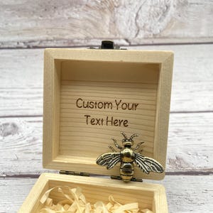 May include: Open wooden box with a hinged lid and a decorative brass bee. The box interior is light-colored wood with the engraved text "Custom Your Text Here". The box is open, revealing shredded paper inside.