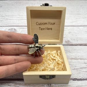 May include: A bronze-colored bee-shaped charm is held in front of an open wooden box. The box is filled with shredded paper and has the text "Custom Your Text Here" engraved on the inside of the lid. The box is closed with a small metal clasp.