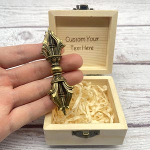 Può includere: Un Vajra color bronzo, un oggetto rituale, è tenuto davanti a una scatola di legno aperta. La scatola è foderata con carta sminuzzata e ha il testo "Custom Your Text Here" inciso all'interno del coperchio. Il Vajra è ornato con intagli dettagliati.