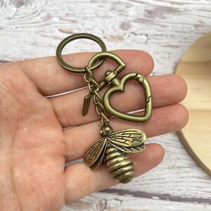 Pu&ograve; includere: Portachiavi color ottone antico con un ciondolo a forma di ape dettagliato. Il portachiavi include un moschettone a forma di cuore e un ciondolo a forma di chiave, collegati da piccole catene e un anello grande.