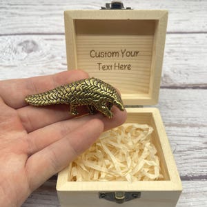 May include: A detailed brass pangolin figurine rests in an open wooden box with the text "Custom Your Text Here" engraved inside. The box is filled with shredded paper. The pangolin is a golden color.