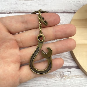 Könnte beinhalten: Antiker, messingfarbener Schlüsselanhänger in Form eines Schraubenschlüssels. Der Schlüsselanhänger hat ein Schraubenschlüssel-Design mit Hakenform und einem kleinen Buchstaben "G"-Anhänger. Das Metall hat ein Vintage-Aussehen. Der Schlüsselanhänger ist an einem Metallring befestigt.