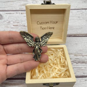 May include: A bronze-colored angel figurine with outstretched wings, held in front of an open wooden box. The box is light-colored with a hinged lid and contains shredded paper. The inside of the lid has the text "Custom Your Text Here".