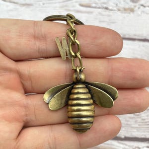 Pu&ograve; includere: Un portachiavi a forma di ape color bronzo con un ciondolo a forma di lettera "W". L'ape ha ali dettagliate e un corpo segmentato. Il portachiavi &egrave; attaccato a un anello e una catena di metallo.