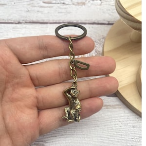 Pode incluir: Um chaveiro de macaco bronzeado. O macaco está sentado, com uma mão na cabeça. O chaveiro tem um anel e uma corrente com um pequeno pingente de letra "D". O fundo é uma superfície de madeira clara.