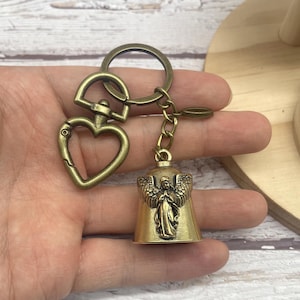 May include: Antique brass-toned keychain featuring a heart-shaped clasp and a bell charm with an angel design. The keychain includes a split ring and a small chain connecting the bell. The overall design is vintage and detailed.
