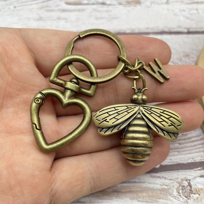 Pu&ograve; includere: Portachiavi color ottone antico con un ciondolo a forma di ape dettagliata con ali spiegate. Il portachiavi include un fermaglio a forma di cuore e un piccolo ciondolo con la lettera "W", il tutto collegato da un portachiavi rotondo. Il ciondolo ape &egrave; lungo circa 3,8 cm.