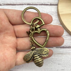 Pu&ograve; includere: Portachiavi color ottone antico con un ciondolo a forma di ape. Il portachiavi include un anello rotondo, un fermaglio a forma di cuore e un ciondolo a forma di ape dettagliato. L'ape ha ali e corpo strutturati.