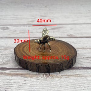 May include: A small, bronze-colored bee figurine, approximately 1.6 inches long and 1.2 inches wide, rests on a round wooden slice. The bee's wings are detailed, and the piece weighs approximately 0.5 ounces. The image includes measurements in millimeters.