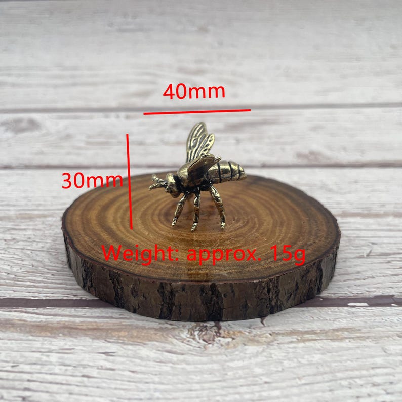 May include: A small, bronze-colored bee figurine, approximately 1.6 inches long and 1.2 inches wide, rests on a round wooden slice. The bee's wings are detailed, and the piece weighs approximately 0.5 ounces. The image includes measurements in millimeters.