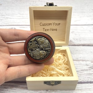 Könnte beinhalten: Eine runde Holzbox mit Scharnierdeckel, geöffnet, um eine dekorative Holzmünze mit einem Bronze-Frosch-Design zu enthüllen. Die Box ist mit dem Text "Custom Your Text Here" graviert. Die Münze wird in einer Hand gehalten.