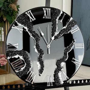 Può includere: Un orologio da parete nero e argento con numeri romani. Il quadrante è nero con lancette e numeri argentati. L'orologio ha un design unico con accenti strutturati grigio scuro. L'orologio misura circa 30 cm di diametro.