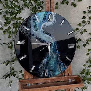 Puede incluir: Un reloj de pared redondo con esfera negra y números romanos plateados. La esfera presenta un diseño abstracto azul y blanco que se asemeja a un río. El reloj se exhibe en un caballete de madera.