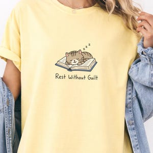 Może przedstawiać: Jasnożółty t-shirt z grafiką brązowego kota pręgowanego śpiącego na otwartej książce oraz napisem "Rest Without Guilt". Kot ma zamknięte oczy, a nad jego głową unoszą się małe czarne symbole "zzz".
