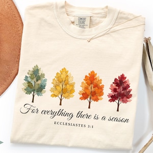 May include: An ivory t-shirt featuring four watercolor-style trees in shades of green, yellow, orange, and red. Below the trees, the text reads, "For everything there is a season" followed by "ECCLESIASTES 3:1" in a black serif and script font.