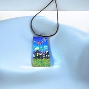 Könnte beinhalten: Eine rechteckige Anhänger-Halskette mit einer Feuerwerksszene. Der Anhänger zeigt einen blauen Himmel mit farbenfrohen Feuerwerken über einer Stadtlandschaft. Der Anhänger ist an einer braunen Kordelkette befestigt.