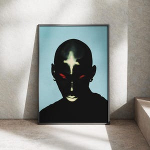 Könnte beinhalten: Gerahmter Kunstdruck mit einem stilisierten Porträt einer Person mit dunkler Haut und rasiertem Kopf. Die Person hat rote Augen und ein weißes Kreuz auf der Stirn. Der Hintergrund ist hellblau.