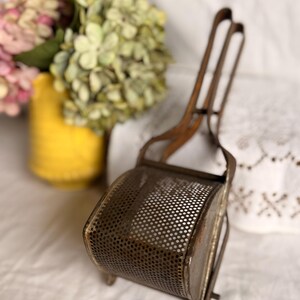 May include: An antique metal potato ricer with a perforated basket and long handles. The ricer is made of a dark, aged metal, and sits on a white surface. A yellow vase with pink and green flowers is in the background.