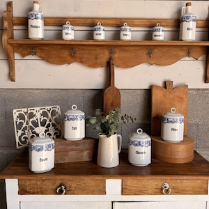 May include: A collection of white ceramic kitchen canisters and bottles featuring blue floral patterns and black text labels including Vinaigre, Epices, Girofle, Poivre, Muscade, Sucre, Cafe, Chicoree, and Pates, arranged on a wooden shelf and cabinet.