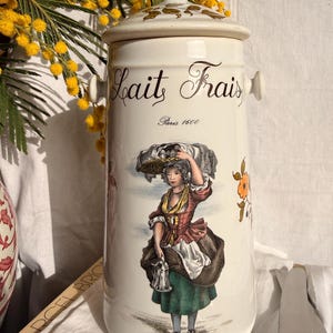Peut inclure: Un grand pot en céramique blanc avec un couvercle, portant les mots "Lait Frais" et "Paris 1600" en écriture noire. Le pot est décoré d'une illustration colorée d'une femme et d'éléments floraux. Le pot a une poignée de chaque côté.