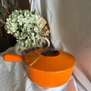 May include: Bright orange enameled cast iron pot with a lid and a long handle. The pot is round and sits on a white surface. The lid has a black handle. A vase of flowers is in the background.