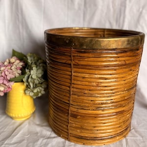 Op de afbeelding: Een cilindrische geweven mand met een messing rand. De mand is gemaakt van natuurlijk gekleurd riet en heeft een horizontaal geribbeld ontwerp. Links van de mand staat een gele vaas met roze en groene bloemen.