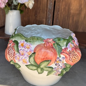 Op de afbeelding: Een witte keramische kom met een verhoogd, handgeschilderd reliëfontwerp van oranje granaatappels, groene bladeren en kleine paarse bloemen. De kom heeft een brede, geschulpte rand en een gladde, glanzende afwerking met botanische details.