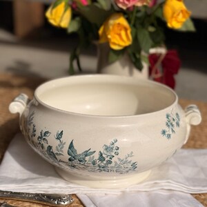 May include: Cream ceramic serving bowl with a teal bird and floral design. The bowl has two handles and a pedestal base. A bouquet of yellow and pink roses is in the background.