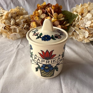 Puede incluir: Tarro de cerámica crema para pimentón con tapa. Decorado con un tulipán rojo, flores azules y la palabra "PAPRIKA" en una pancarta. La tapa tiene un pequeño asa y diseños florales. Flores beige en el fondo.
